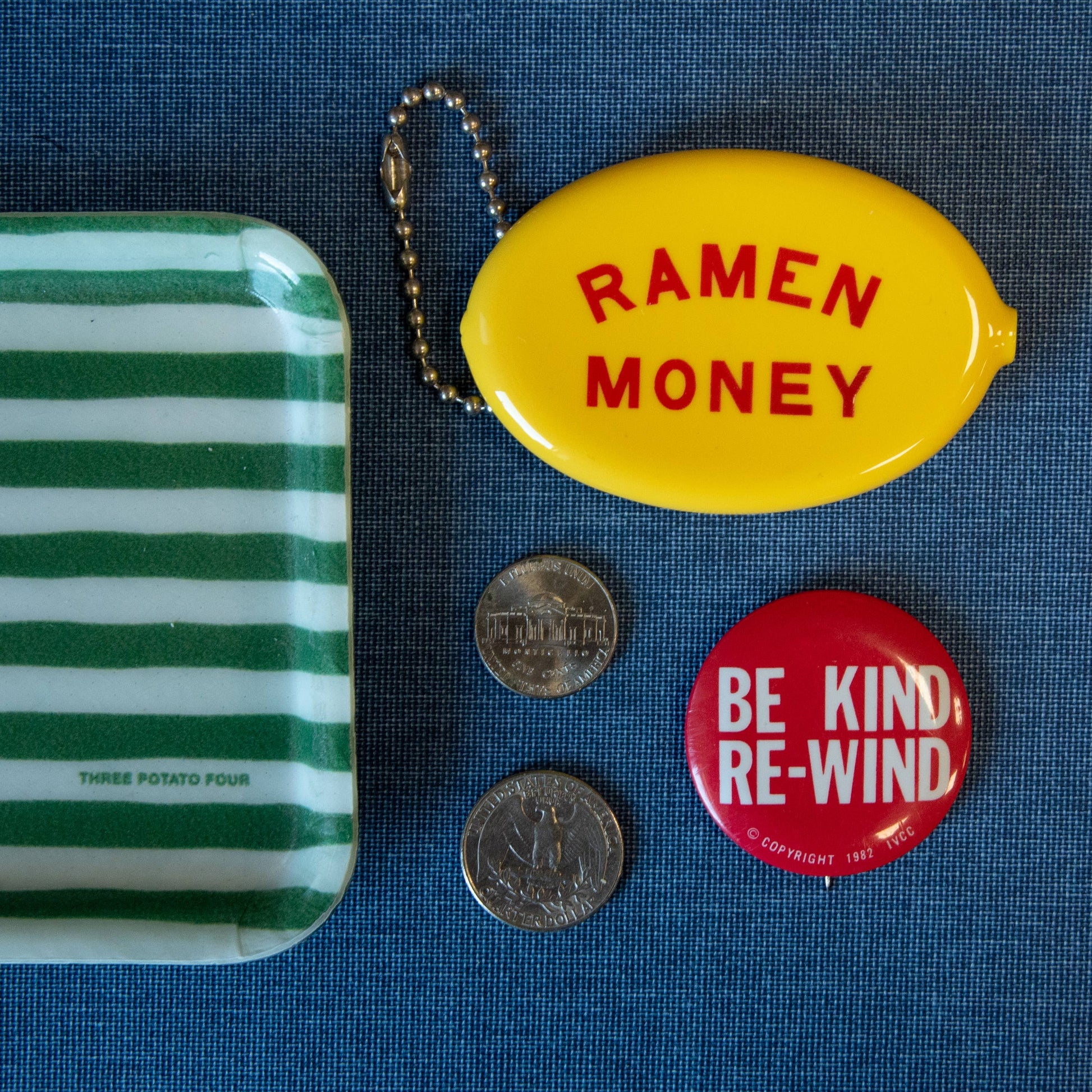Collection of items including a striped tin, yellow 'Ramen Money' container, coins, and a 'Be Kind Re-Wind' button on a blue background.