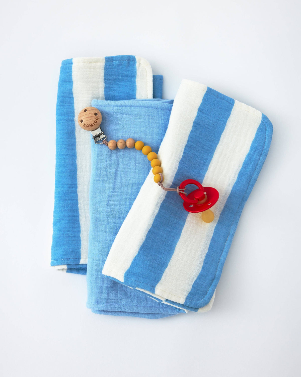 Two folded muslin baby blankets with blue and white stripes, styled with a wooden teething ring and a red pacifier clip on a white background.