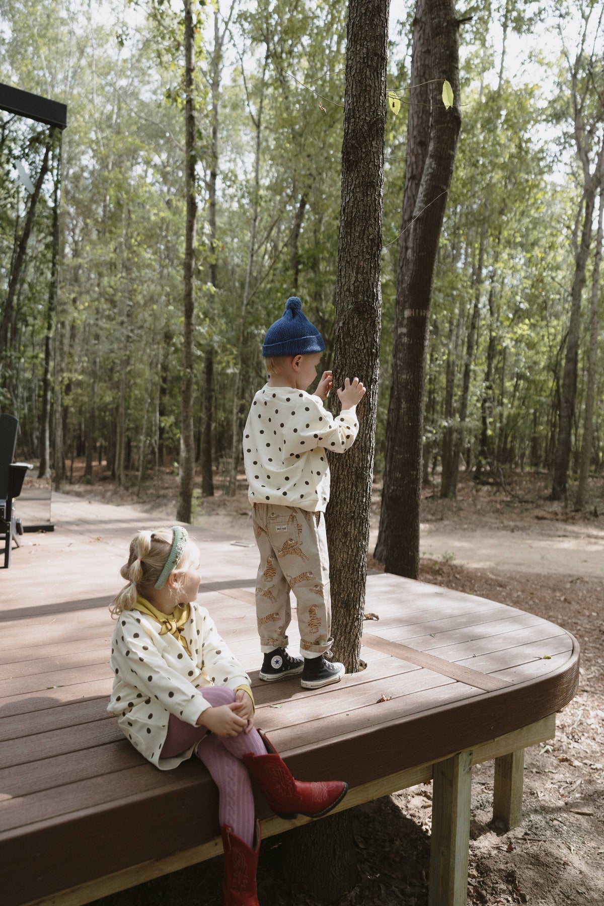 Two children outdoors on a wooden deck in a wooded area. One child stands near a tree wearing a cream polka-dot outfit with a blue beanie, and the other sits on the deck wearing a cream polka-dot top with red boots.