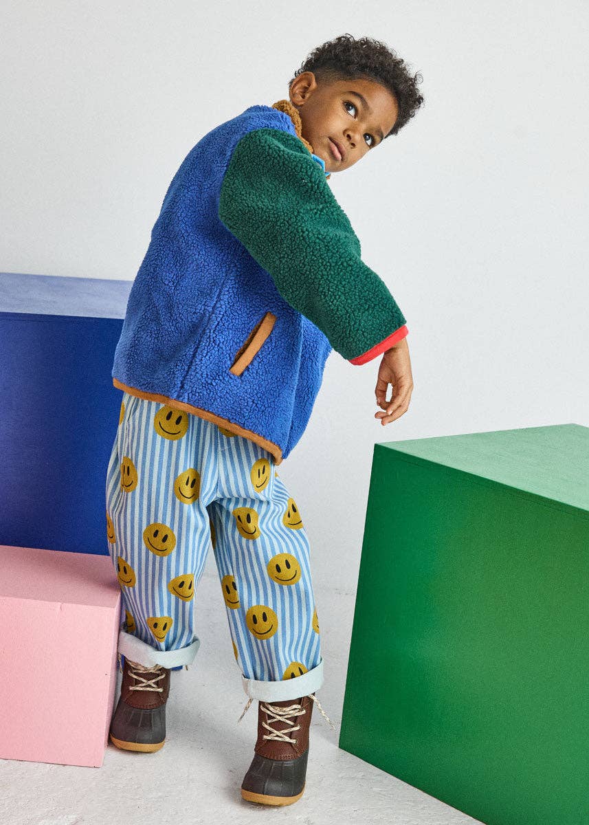 A young child modeling a colorful fleece jacket with blue and green panels, matching striped smiley-face pants, and brown boots, posed in front of geometric color blocks.