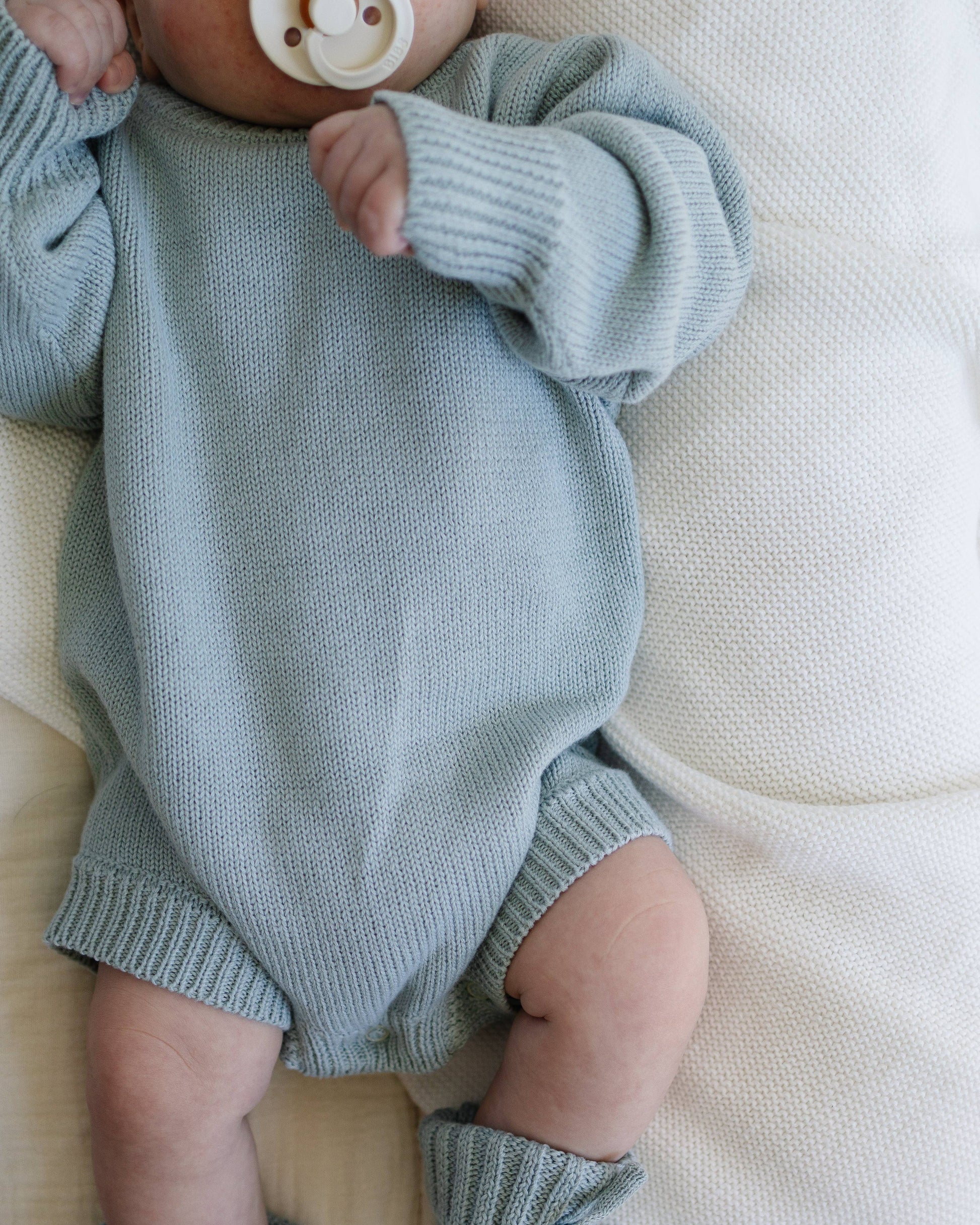 Baby wearing a light blue knitted outfit on a white blanket