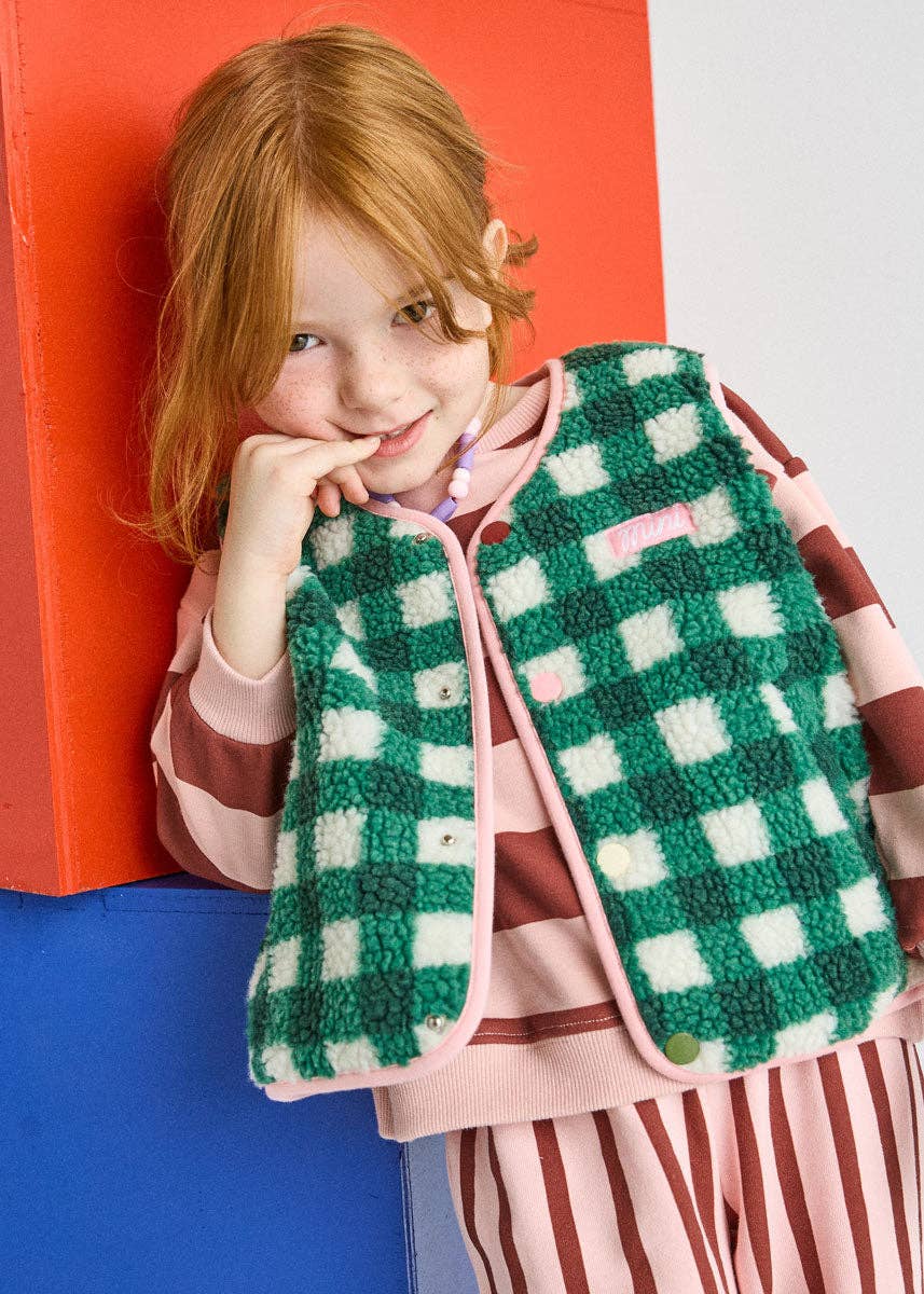 A young girl with red hair leans against a colorful background, wearing a green and white checkered fleece vest over a pink and brown striped long-sleeve top and matching striped pants.
