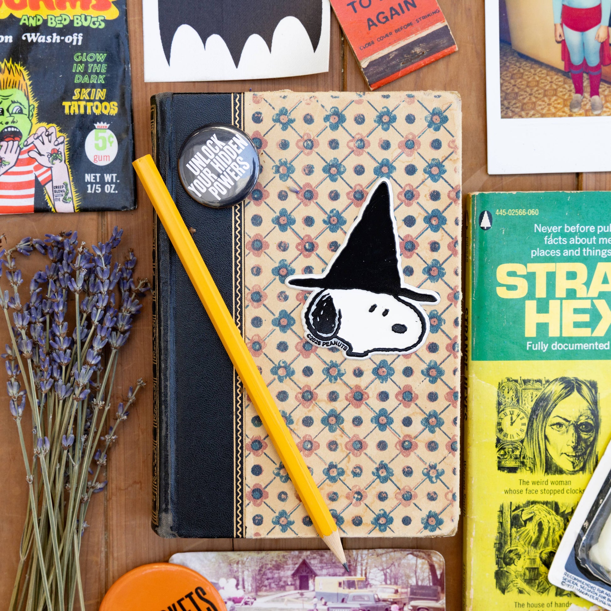 A styled flat-lay featuring a vintage patterned notebook with a Snoopy witch magnet a yellow pencil, a round pin, dried lavender, and retro magazine and book covers arranged on a wooden surface.