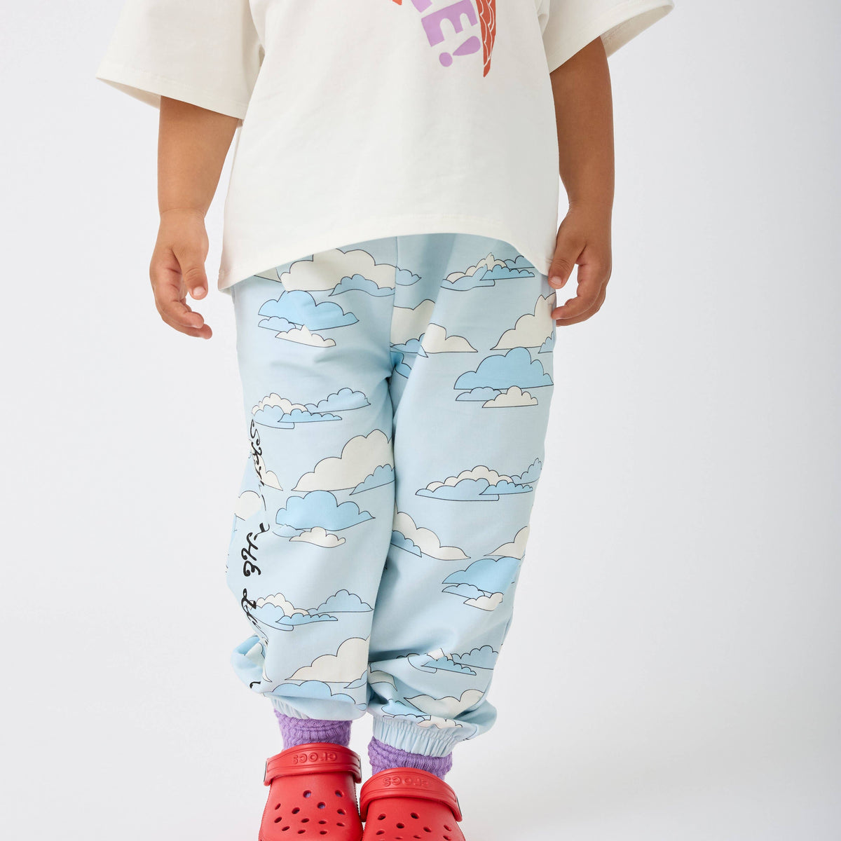 A child wearing light blue jogger-style pants with a cloud print, paired with a cream t-shirt and red clogs, shown from the waist down against a plain white studio background.