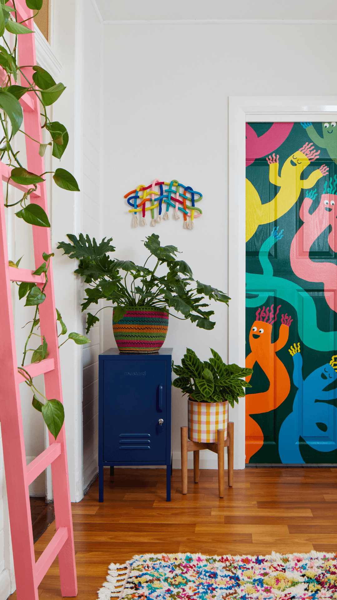 A colorful interior corner with a pink ladder, potted green plants, a small blue cabinet, and a door covered in bright multicolored abstract monster illustrations.